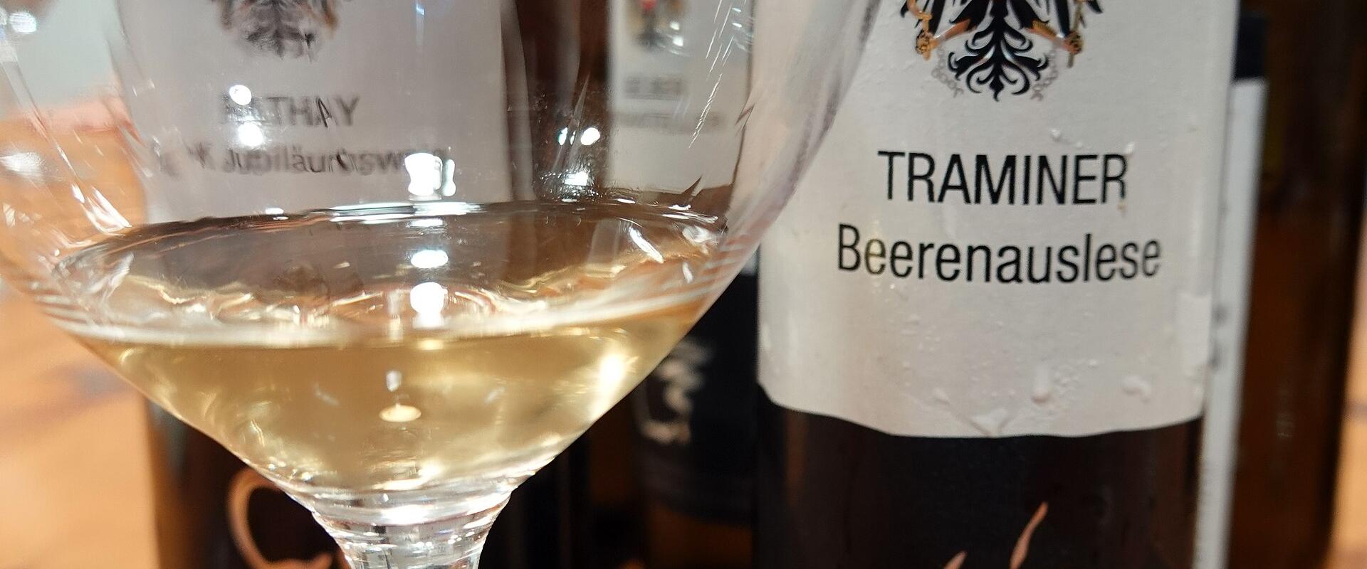 A close-up of wine bottles and a tasting glass on a wooden table, with labels showing the Austrian eagle emblem and text including "TRAMINER Beerenauslese" and "BUNDESLEHRANSTALT UND BUNDESAMT FÜR WEIN- UND OBSTBAU KLOSTERNEUBURG".