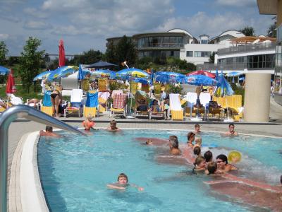 An outdoor swimming pool area with people swimming and relaxing on lounge chairs under umbrellas, with modern resort buildings in the background.