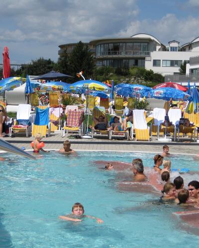 An outdoor swimming pool area with people swimming and relaxing on lounge chairs under umbrellas, with modern resort buildings in the background.