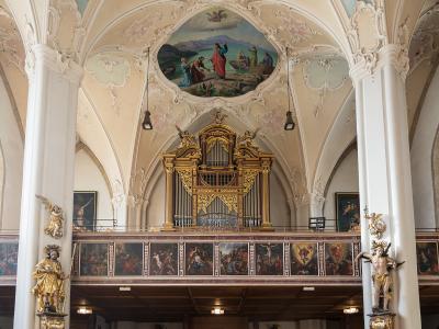 The interior of a Baroque church featuring a large golden pipe organ on a gallery, flanked by gilded statues and a painted ceiling fresco.