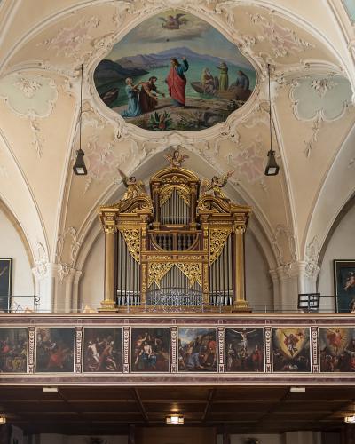 The interior of a Baroque church featuring a large golden pipe organ on a gallery, flanked by gilded statues and a painted ceiling fresco.