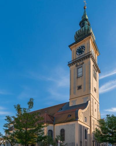 A low-angle photograph of a large Baroque-style church with a tall, pale yellow bell tower topped by a green onion dome and cross, set against a clear blue sky with trees in the foreground.
