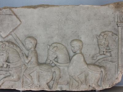 A close-up photograph of a weathered Roman stone relief depicting two figures on horseback, with one figure holding a large rectangular shield, displayed against a plain wall.
