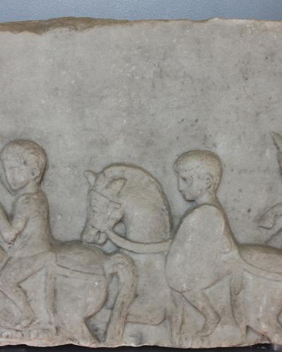 A close-up photograph of a weathered Roman stone relief depicting two figures on horseback, with one figure holding a large rectangular shield, displayed against a plain wall.