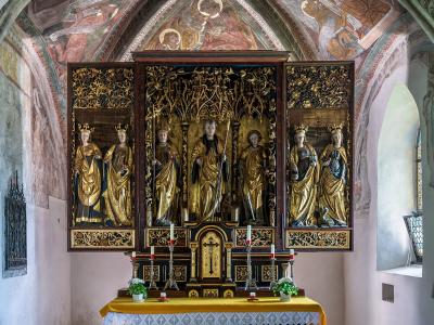 The image shows a gilded wooden altarpiece with multiple carved figures, situated inside a chapel with frescoed walls and vaulted ceilings.