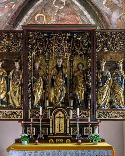 The image shows a gilded wooden altarpiece with multiple carved figures, situated inside a chapel with frescoed walls and vaulted ceilings.