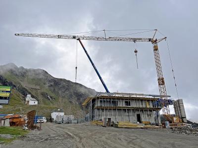 A construction site in a mountainous region featuring a large yellow tower crane, a blue mobile crane, and a multi-story concrete building under construction, with a green directional sign on the left listing "Gletscher," "Rifflsee," and "Ködlsee."
