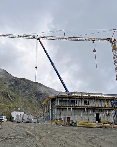 A construction site in a mountainous region featuring a large yellow tower crane, a blue mobile crane, and a multi-story concrete building under construction, with a green directional sign on the left listing "Gletscher," "Rifflsee," and "Ködlsee."