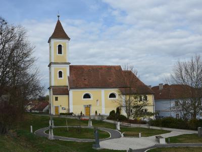 A yellow church with a tall, pointed tower and a red-tiled roof, situated on a grassy hill with a paved circular path and statues in the foreground.