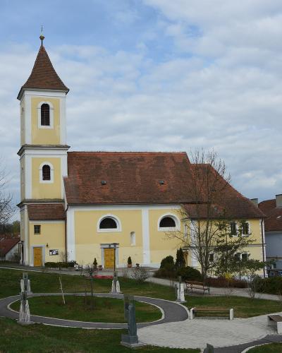 A yellow church with a tall, pointed tower and a red-tiled roof, situated on a grassy hill with a paved circular path and statues in the foreground.