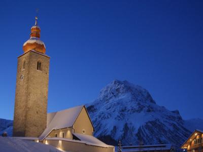 A stone church with a distinctive onion dome tower illuminated at dusk, set against a snow-covered mountain backdrop, with a sign for "TIEFGARAGE ANGER" visible in the foreground.
