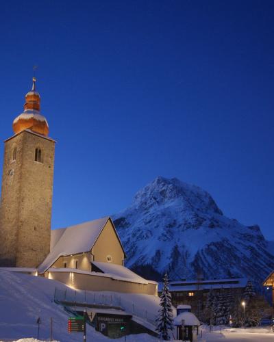 A stone church with a distinctive onion dome tower illuminated at dusk, set against a snow-covered mountain backdrop, with a sign for "TIEFGARAGE ANGER" visible in the foreground.