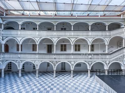 The image shows a multi-story interior courtyard (arkadenhof) with white arcades, balustrades, and a checkered floor, covered by a modern glass roof structure.