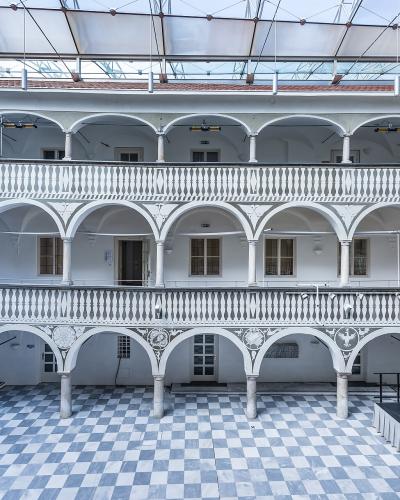 The image shows a multi-story interior courtyard (arkadenhof) with white arcades, balustrades, and a checkered floor, covered by a modern glass roof structure.