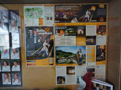 A corkboard display featuring a large poster for "Salzwelten Salzburg" alongside a map of the Hallein salt mine, informational brochures, and a grid of staff photos.