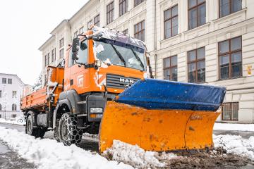 An orange MAN TGA dump truck equipped with a large snowplow blade is clearing snow from a city street, with the word "Leoben" visible on the truck's door and a multi-story building in the background.