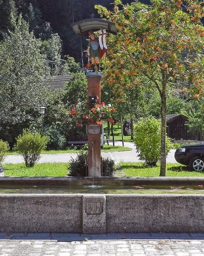 A stone fountain with a central wooden post featuring carved figures and flowers, situated in a paved area with parked cars and traditional alpine buildings in the background.