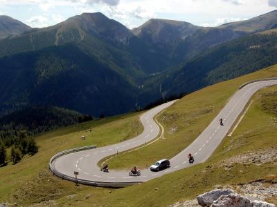 A winding mountain road with hairpin turns traversing a grassy alpine slope, featuring a car and several motorcycles, with forested mountains in the background.