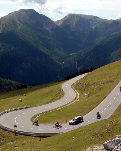 A winding mountain road with hairpin turns traversing a grassy alpine slope, featuring a car and several motorcycles, with forested mountains in the background.
