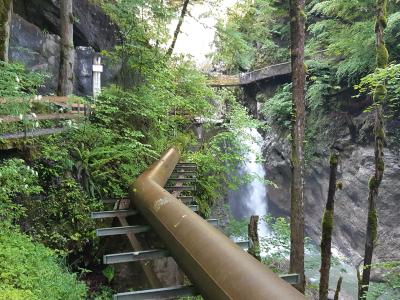 A view inside a narrow, rocky gorge featuring a large industrial pipe running alongside a wooden staircase and walkway, with a waterfall in the background.