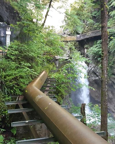 A view inside a narrow, rocky gorge featuring a large industrial pipe running alongside a wooden staircase and walkway, with a waterfall in the background.