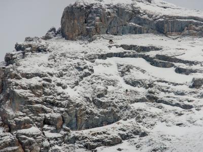 A snowy, rocky mountain slope with a small structure and a person visible near the top.