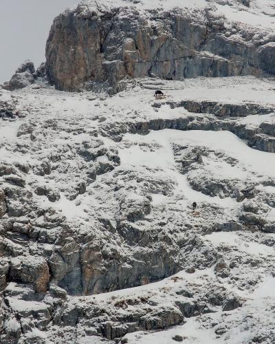 A snowy, rocky mountain slope with a small structure and a person visible near the top.