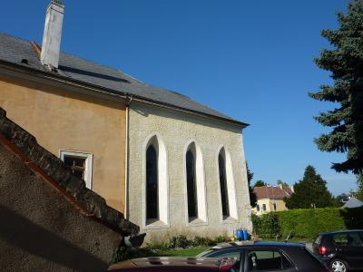 A low-angle shot of a building with a yellow stucco wall and three tall, narrow Gothic-style arched windows on a textured white section, with a stone wall in the foreground and cars parked nearby.