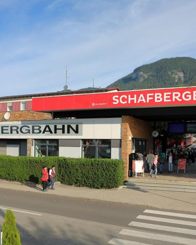 The image shows the exterior of a modern building with a large red sign reading "SCHAFBERGBAHN" and a smaller sign below it also reading "SCHAFBERGBAHN", with people gathered near the entrance and a mountain in the background.