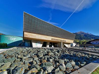 A modern visitor center building with a distinctive slanted wooden facade and a large green cylindrical structure labeled "Nationalpark 360°" set against a mountain backdrop.