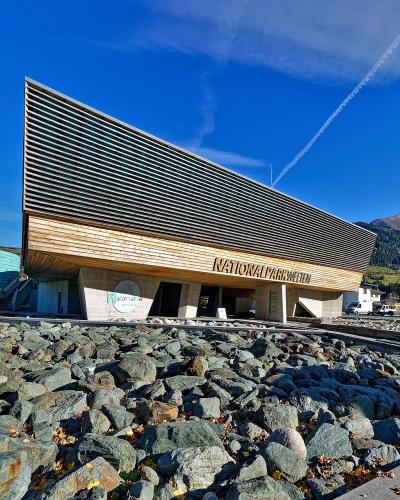 A modern visitor center building with a distinctive slanted wooden facade and a large green cylindrical structure labeled "Nationalpark 360°" set against a mountain backdrop.