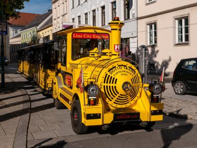 A yellow tourist train vehicle labeled "Linz City Express" and "DOTTO TRAINS CASTELFRANCO V. ITALY" is parked on a paved street lined with multi-story buildings.