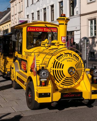 A yellow tourist train vehicle labeled "Linz City Express" and "DOTTO TRAINS CASTELFRANCO V. ITALY" is parked on a paved street lined with multi-story buildings.