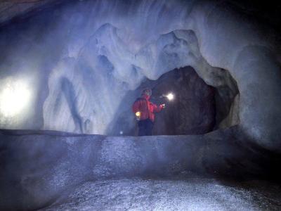 A person in a red jacket and helmet stands inside a large ice cave, holding a flashlight that illuminates the surrounding blue-white ice formations and a dark rock archway.