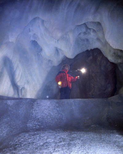 A person in a red jacket and helmet stands inside a large ice cave, holding a flashlight that illuminates the surrounding blue-white ice formations and a dark rock archway.