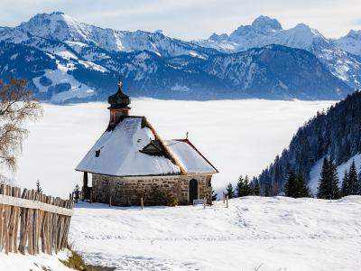 A stone chapel with a dark onion dome sits on a snowy ridge, overlooking a valley filled with a thick layer of fog, with snow-capped mountains in the background.