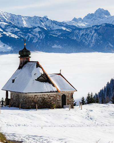 A stone chapel with a dark onion dome sits on a snowy ridge, overlooking a valley filled with a thick layer of fog, with snow-capped mountains in the background.