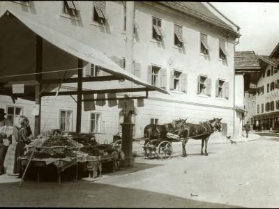 A black and white historical photograph showing a market stall with produce under a large awning on the left, a horse-drawn cart in the center, and a large multi-story building with shuttered windows in the background.