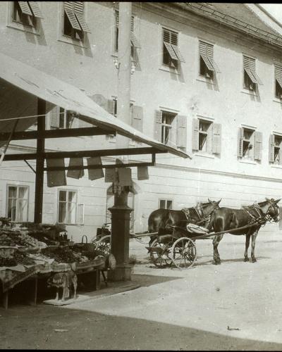 A black and white historical photograph showing a market stall with produce under a large awning on the left, a horse-drawn cart in the center, and a large multi-story building with shuttered windows in the background.