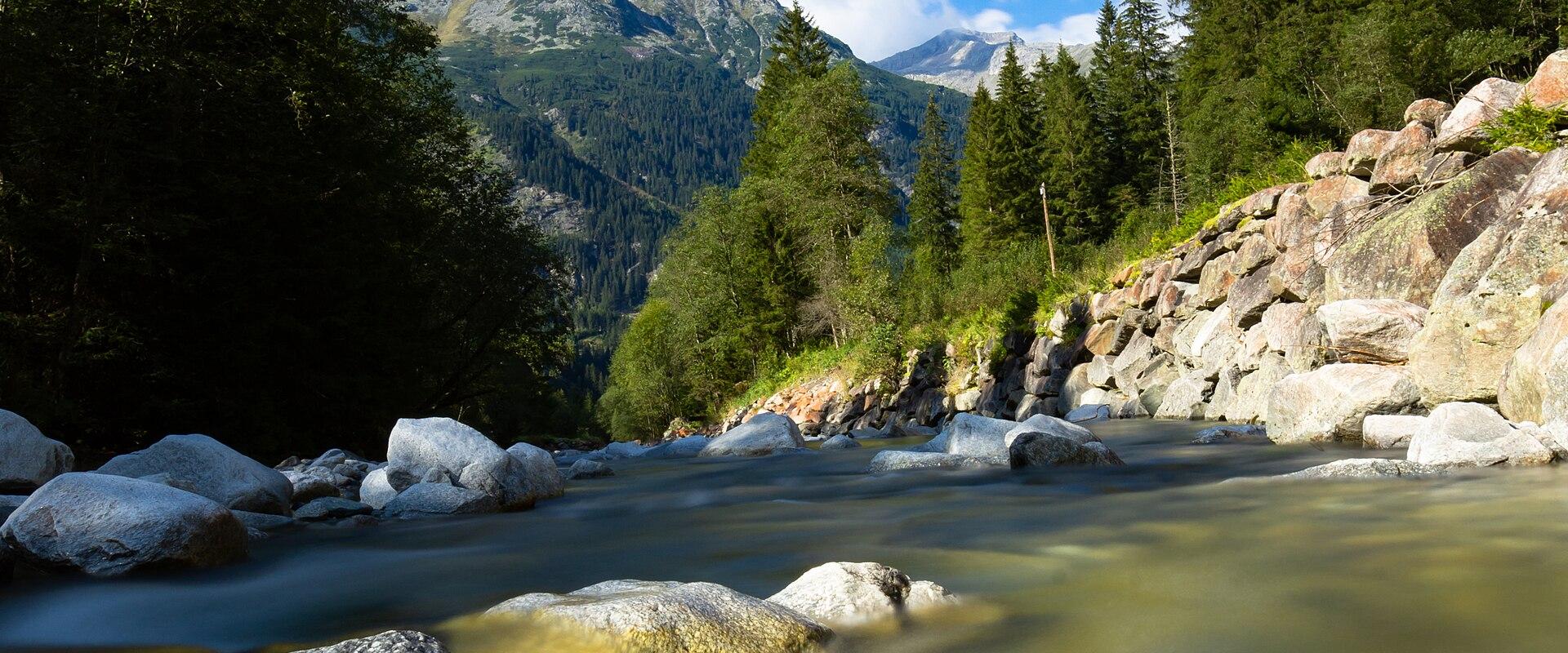 A long-exposure photograph of a glacial river flowing through a rocky, forested mountain valley with snow-capped peaks in the background under a clear blue sky.