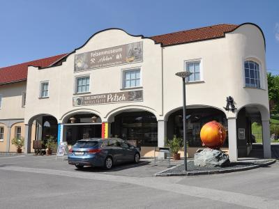 A cream-colored building with a red-tiled roof and arched ground-floor walkways, featuring a large banner reading "Felsenmuseum" and a smaller sign for "Edelserpentin WERKSTÄTTE Patsch," with a blue car parked in front and a large orange sphere sculp