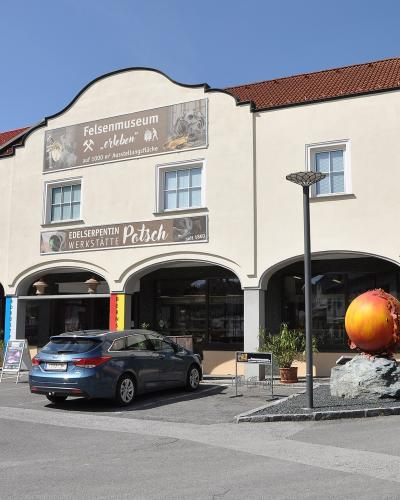 A cream-colored building with a red-tiled roof and arched ground-floor walkways, featuring a large banner reading "Felsenmuseum" and a smaller sign for "Edelserpentin WERKSTÄTTE Patsch," with a blue car parked in front and a large orange sphere sculp