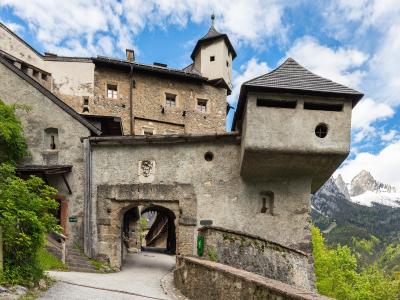 A stone fortress entrance with a prominent overhanging tower, a coat of arms above the archway, and a view of snow-capped mountains in the background.