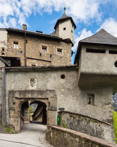 A stone fortress entrance with a prominent overhanging tower, a coat of arms above the archway, and a view of snow-capped mountains in the background.