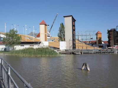 A large outdoor theatre complex built on a lake, featuring tiered yellow seating, distinctive towers with red roofs, and extensive scaffolding, viewed from a wooden walkway.