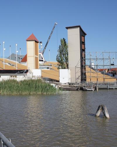 A large outdoor theatre complex built on a lake, featuring tiered yellow seating, distinctive towers with red roofs, and extensive scaffolding, viewed from a wooden walkway.