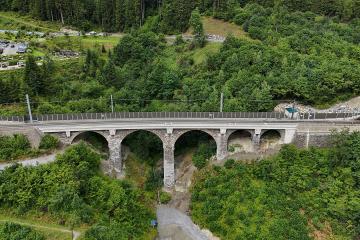Aerial view of a multi-arch stone railway viaduct with overhead catenary wires, set in a densely forested valley with a gravel riverbed below and a parking area visible on the hillside to the left.