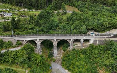 Aerial view of a multi-arch stone railway viaduct with overhead catenary wires, set in a densely forested valley with a gravel riverbed below and a parking area visible on the hillside to the left.