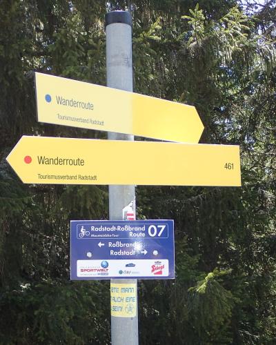 A metal signpost in a forest setting displaying yellow hiking route signs labeled "Wanderroute" with "Tourismusverband Radstadt" and a blue mountain bike route sign for "Radstadt-Roßbrand Route 07" pointing to "Roßbrand" and "Radstadt".