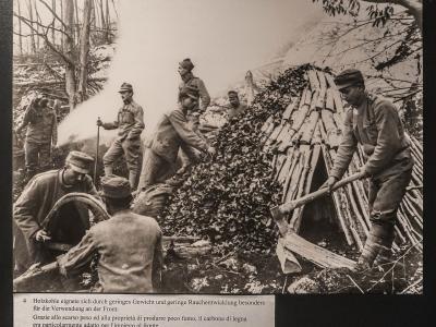 A black and white historical photograph showing soldiers in World War I-era uniforms constructing a charcoal kiln in a forest, with a caption below in German and Italian explaining the utility of charcoal for the front lines.
